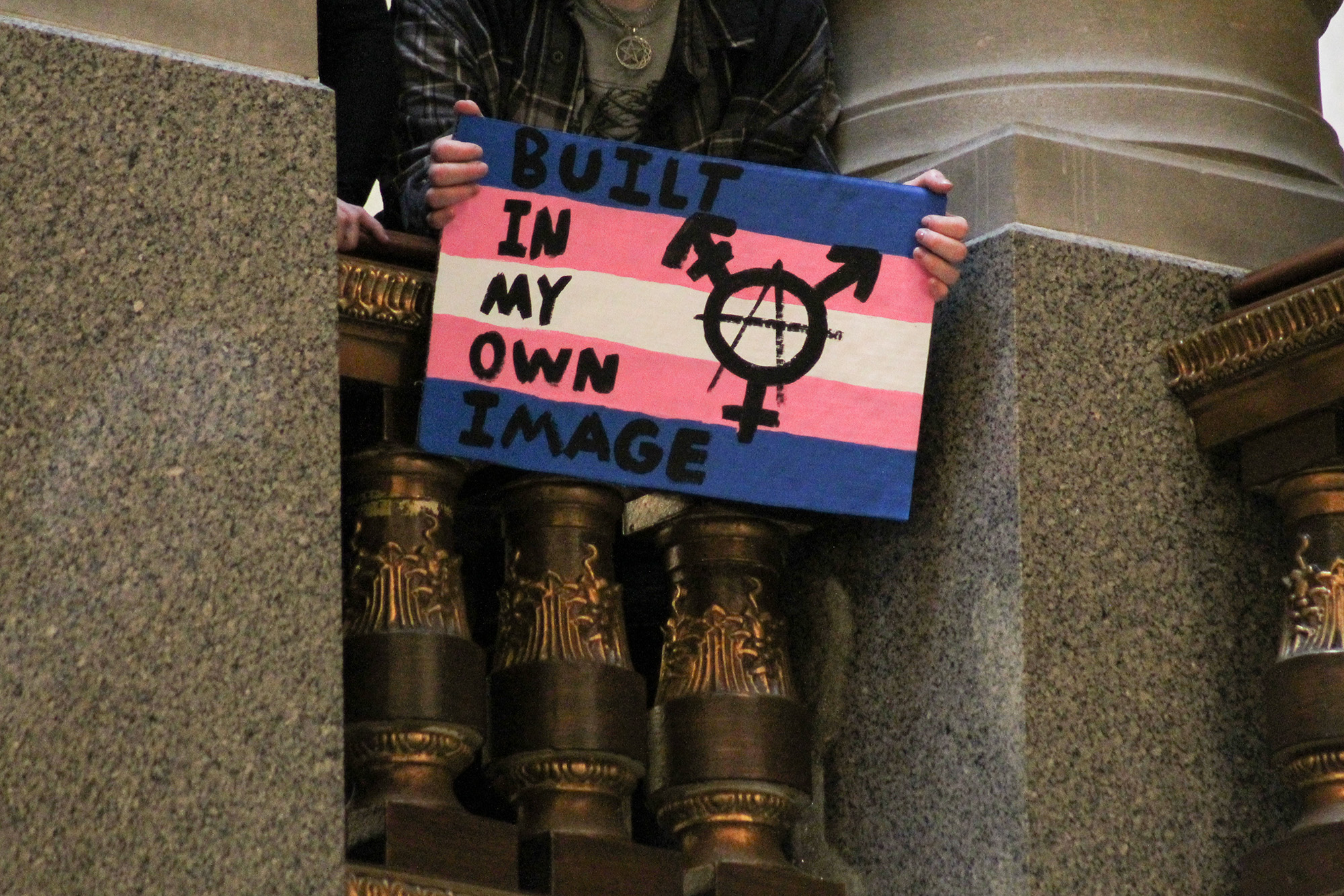 A protester hangs over the edge of the second floor of the Indiana Statehouse. They hold a sign painted in transgender pride flag colors, written on it is 'Built in my own image.'
